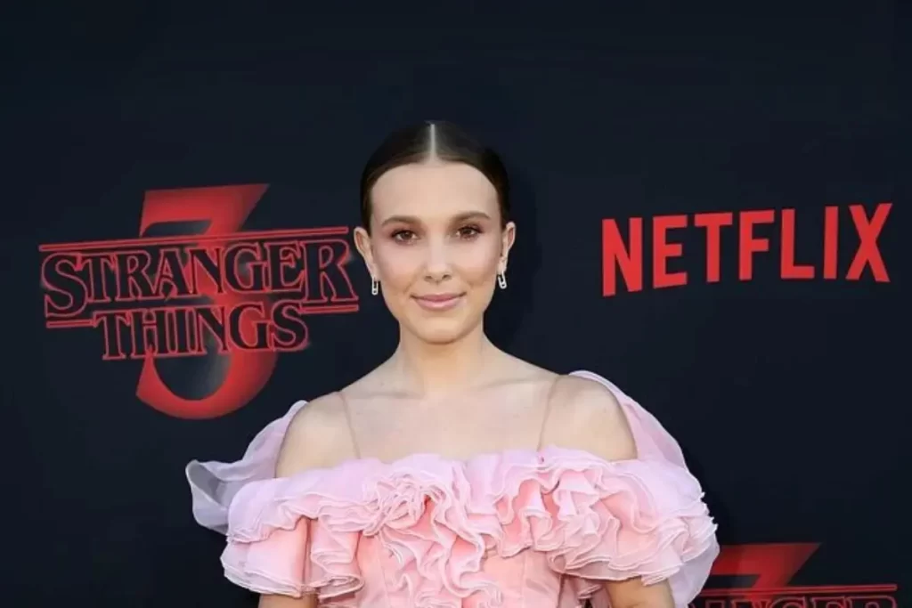 Millie Bobby Brown sorrindo suavemente para a câmera, com um fundo desfocado que sugere um evento de promoção de filme.